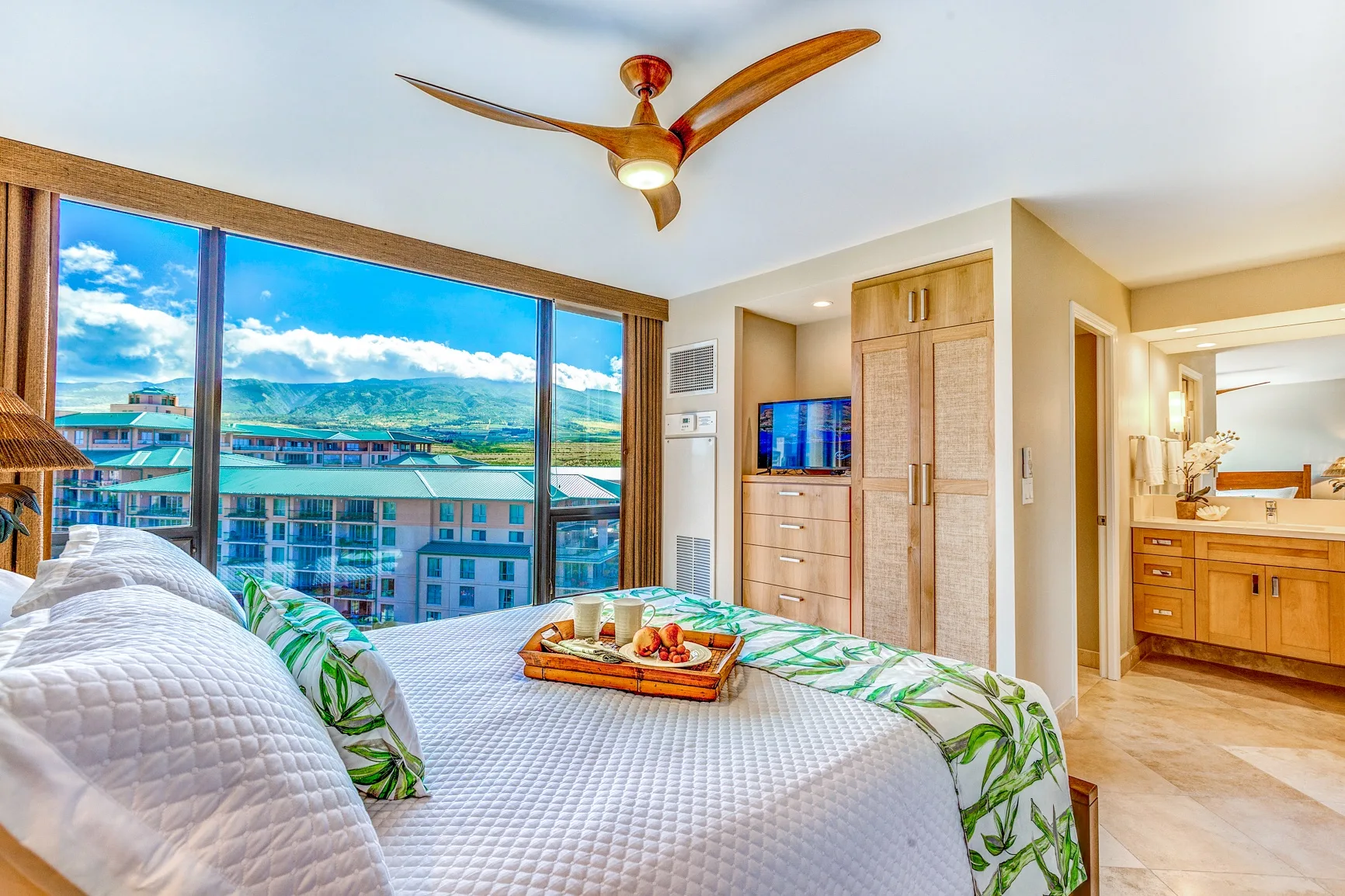 A bedroom with a bed, a television, and a wide window overlooking nearby buildings.