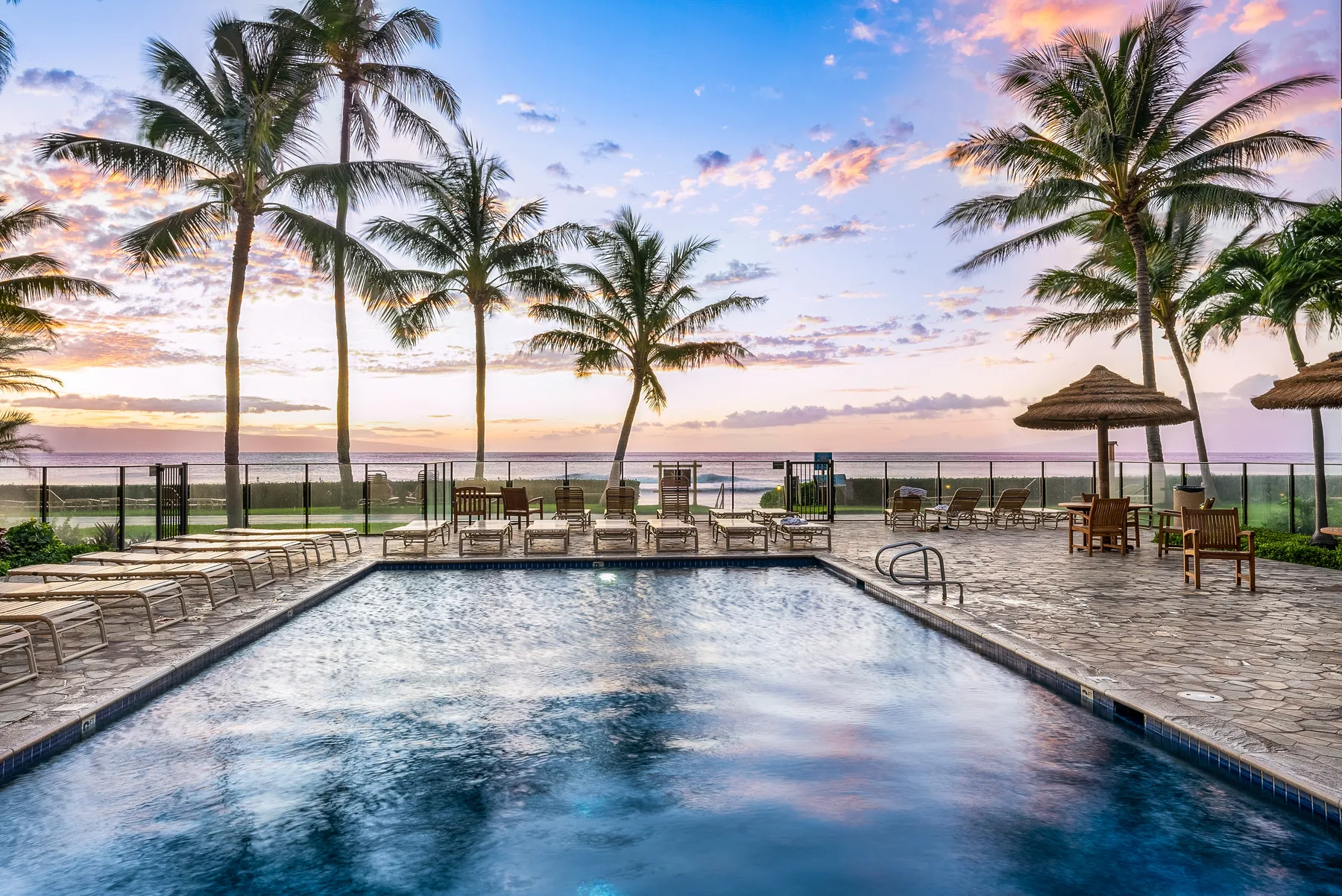A swimming pool faces a beach, with palm trees along the shore at sunset.