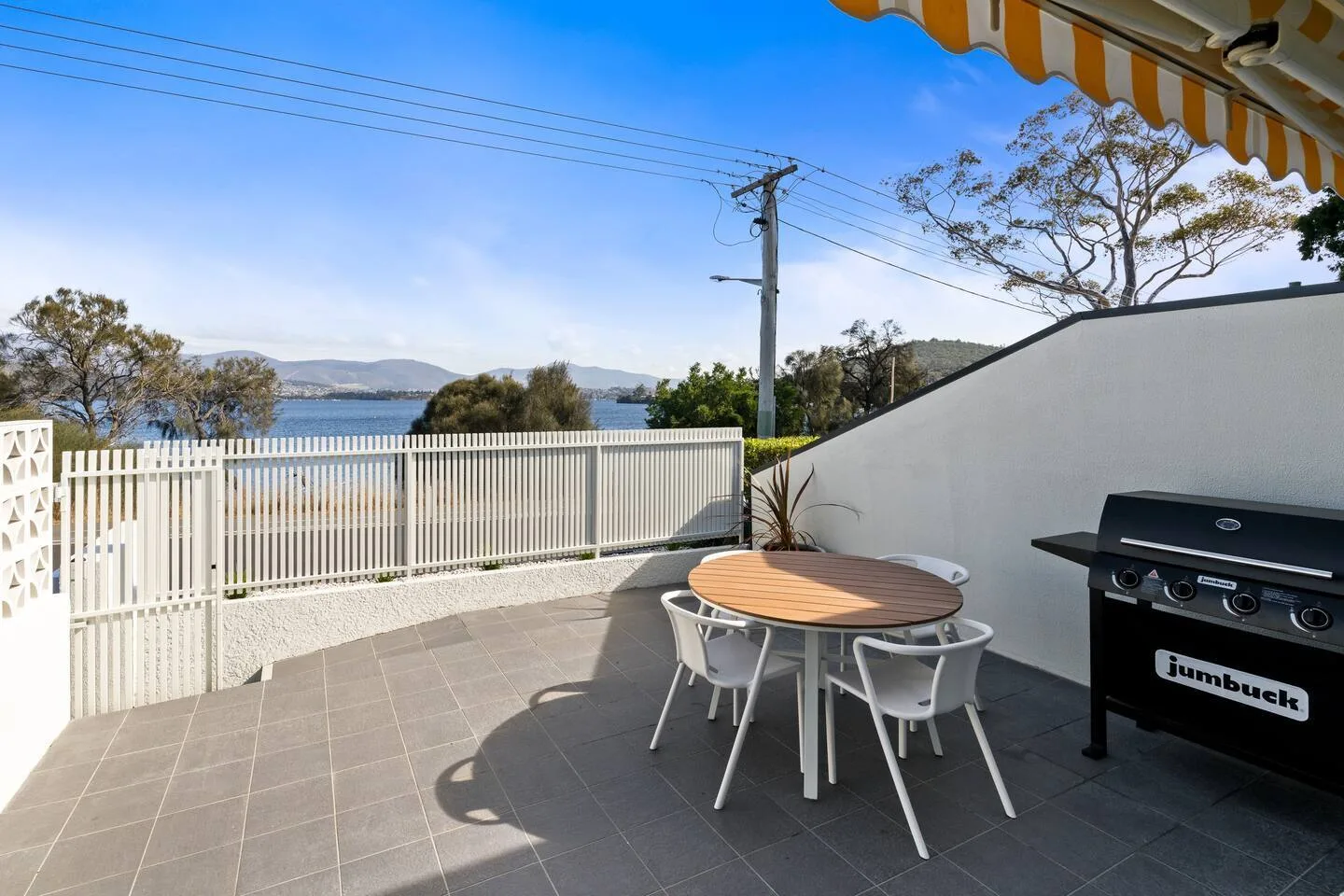 An outdoor patio has a dining table and barbecue beside a white fence overlooking the water.