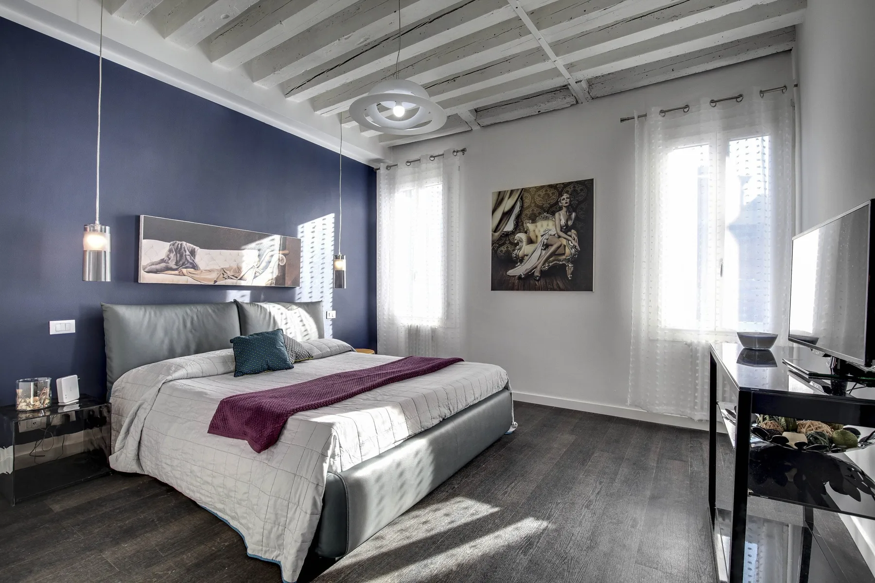 Un dormitorio con una cama, una pared azul oscura y ventanas altas con cortinas.