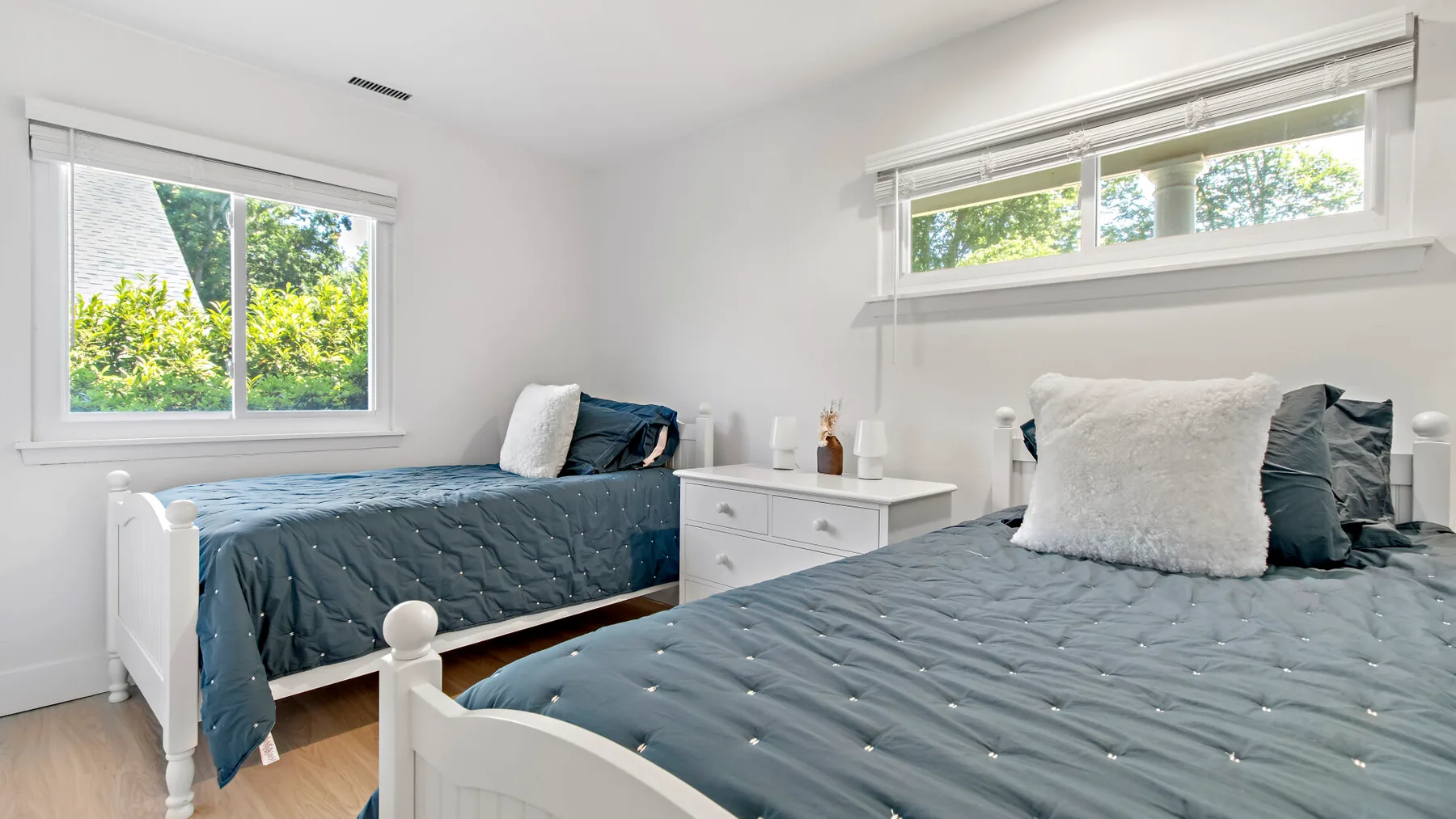 A bright bedroom with two beds dressed in blue bedding and windows overlooking greenery.