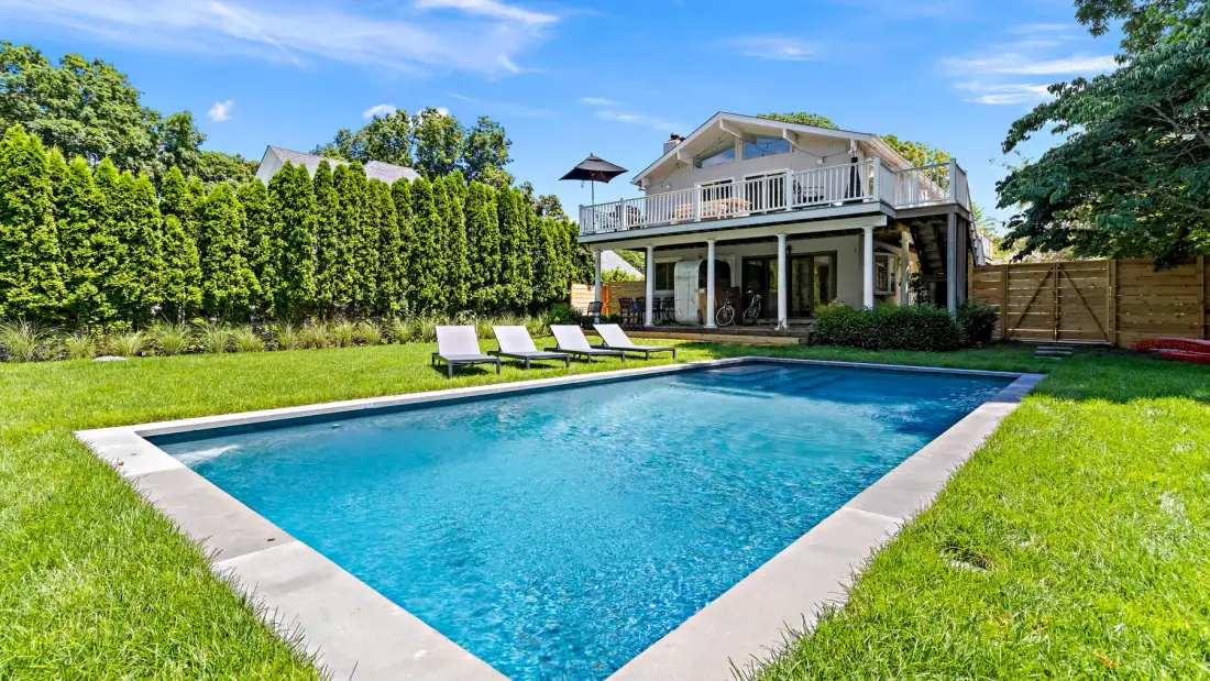 A backyard swimming pool sits beside a two-story house and green lawn.