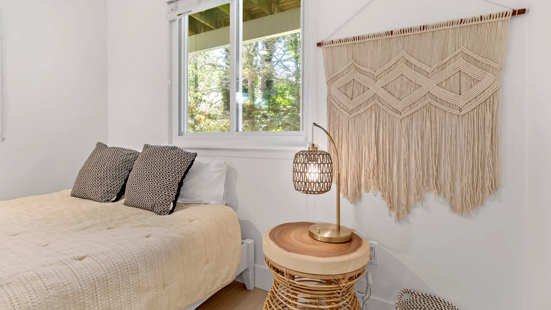 A bright bedroom with a bed, patterned pillows, and a window overlooking greenery.