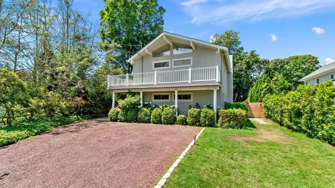 A raised house with a long balcony stands beside a broad driveway and green lawn.