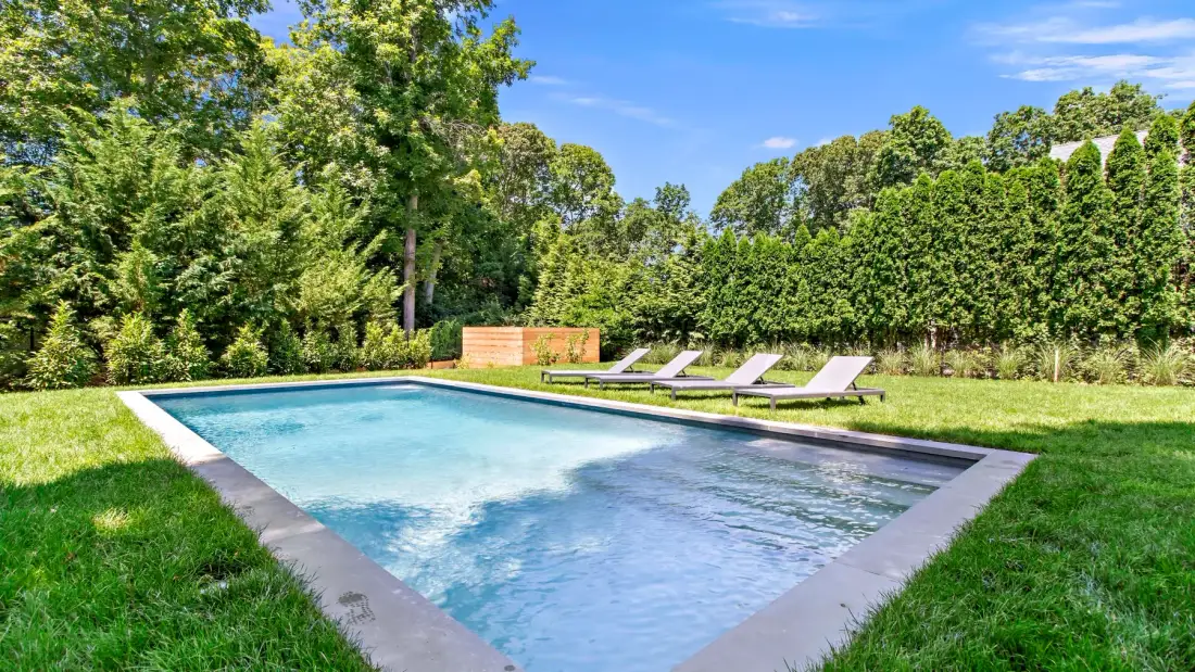 An outdoor swimming pool sits in a grassy yard bordered by trees and hedges.