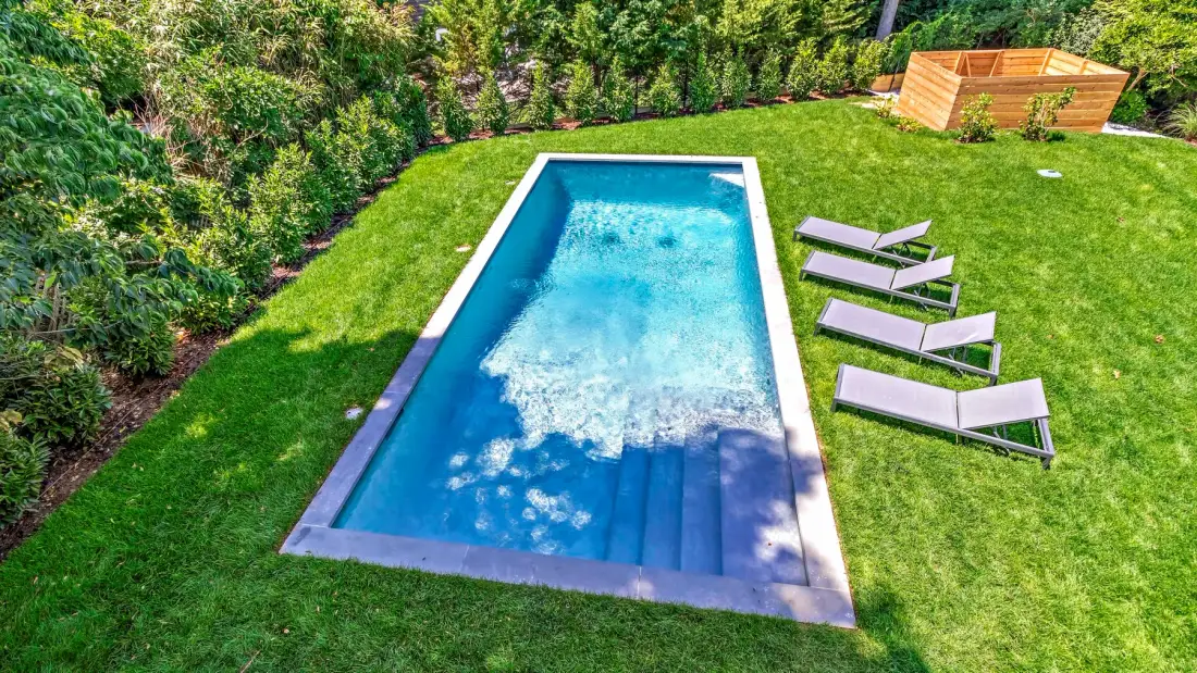 A rectangular swimming pool sits in a green lawn beside several lounge chairs.