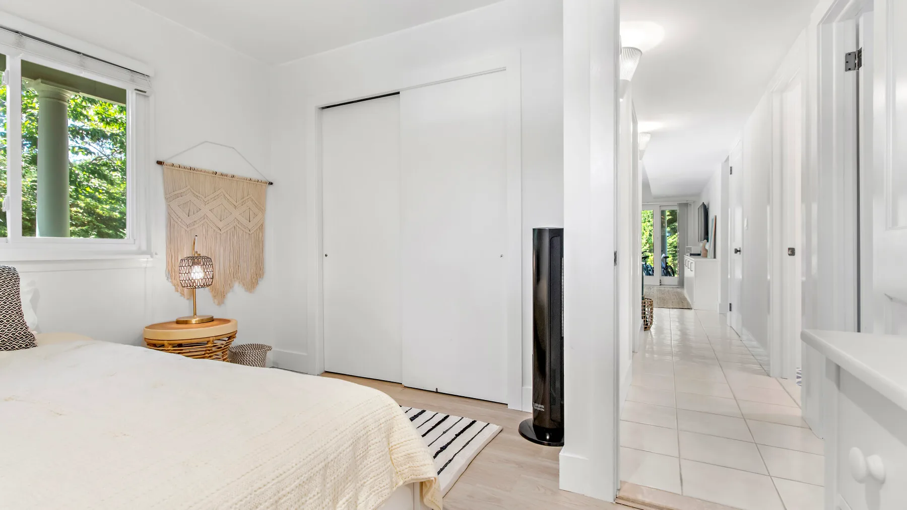 A bright bedroom with a bed, window, and a hallway visible through the doorway.