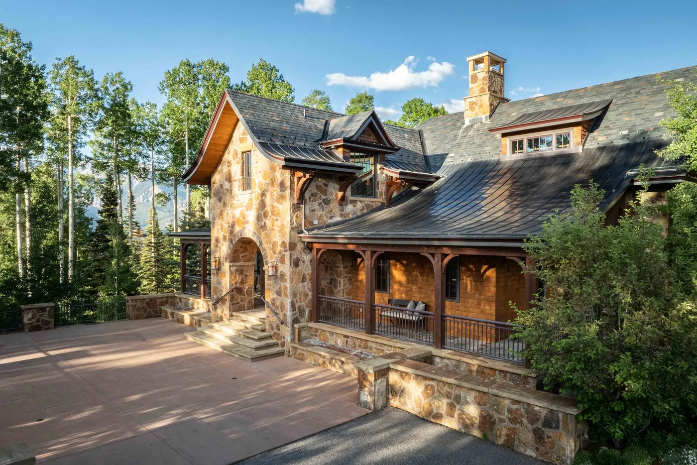 A large stone house with a covered porch sits beside a wooded landscape.