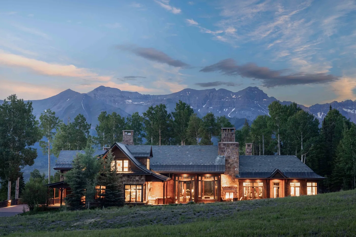 A warmly lit lodge sits beneath a mountain range at dusk.