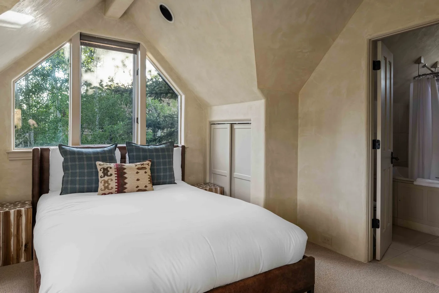 A bedroom with a bed beneath a large window overlooking leafy trees.