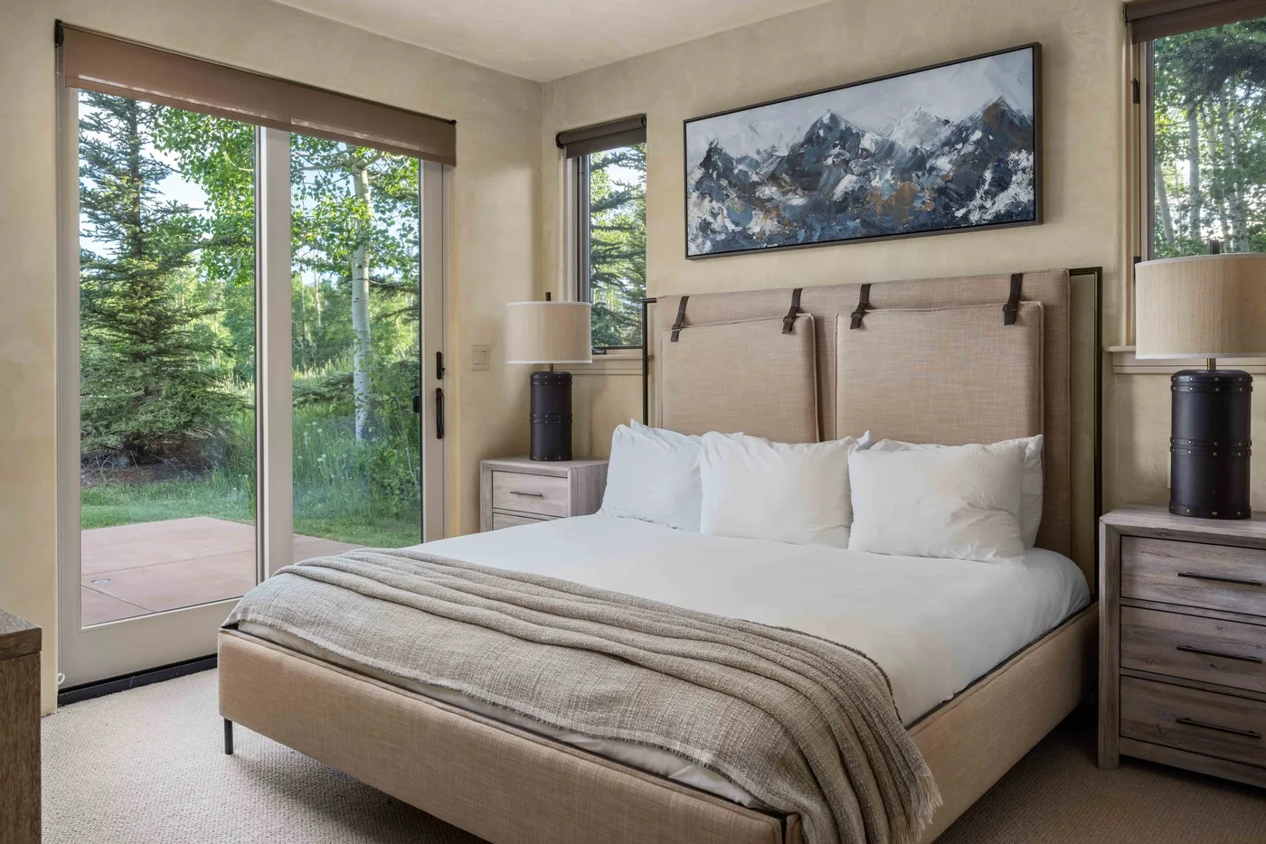 A bedroom with a neatly made bed and large windows overlooking trees.