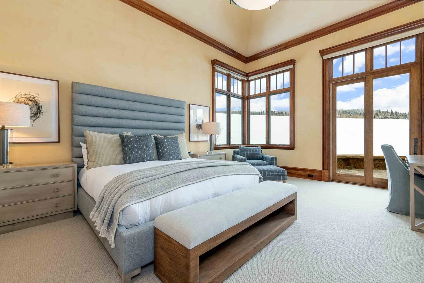 A bright bedroom with a large bed, upholstered bench, and tall windows overlooking a snowy landscape.