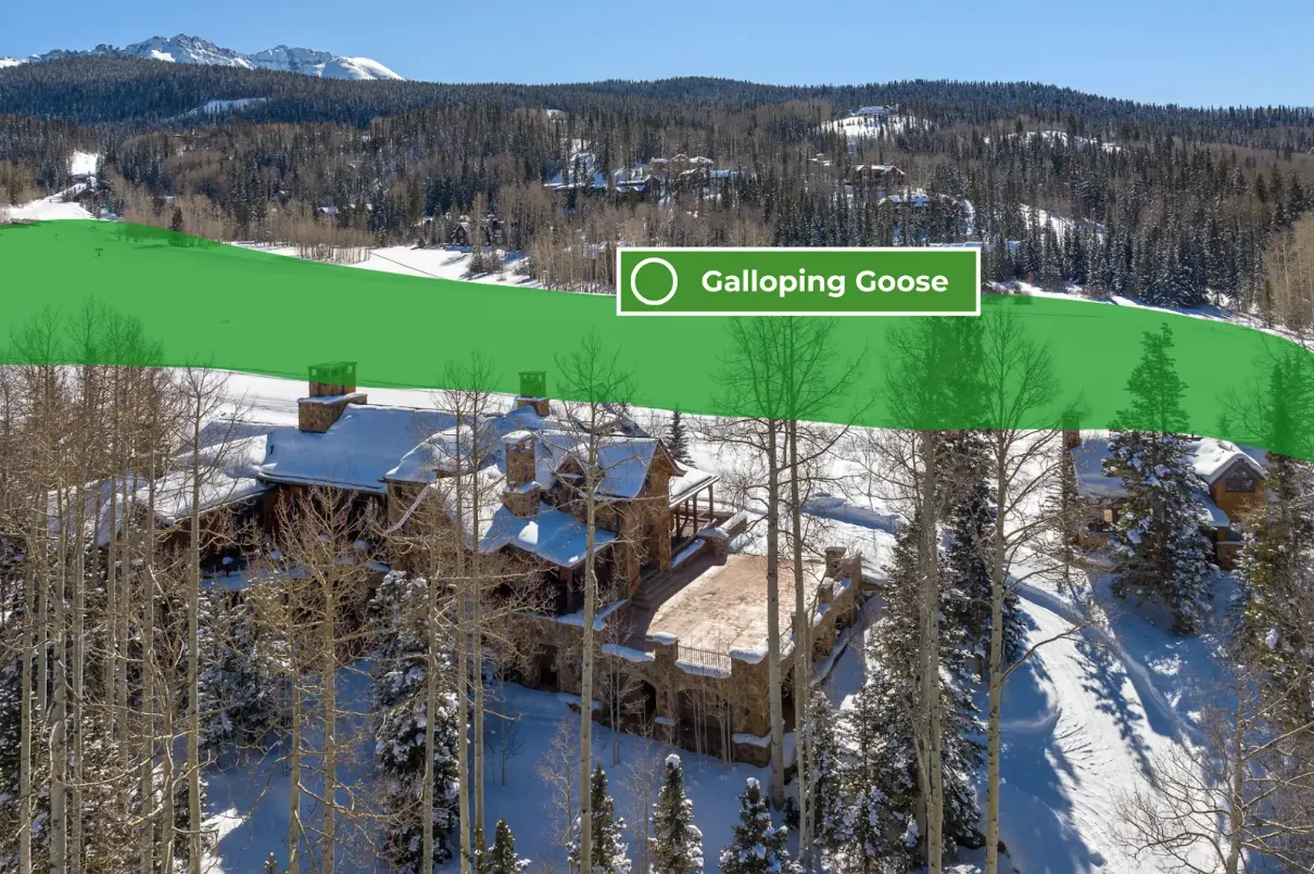 Snow-covered buildings sit among trees on a wooded hillside beneath distant mountains.