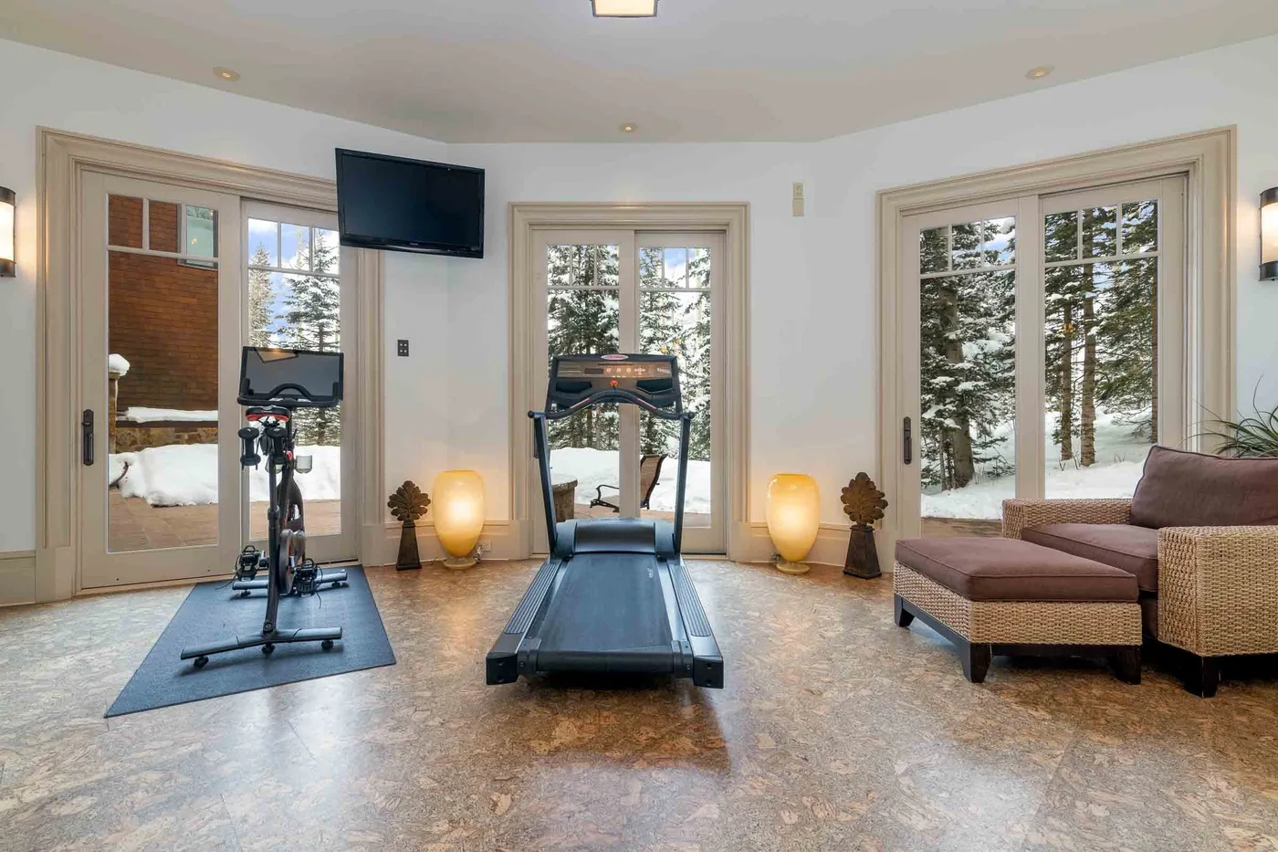 A bright exercise room with a treadmill and stationary bike beside large windows.