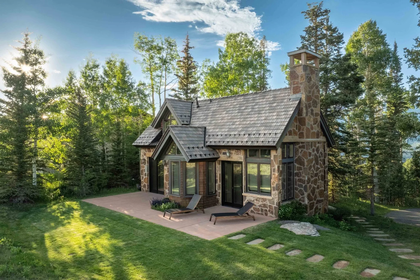 A stone-and-wood cabin stands beside a patio in a wooded setting.