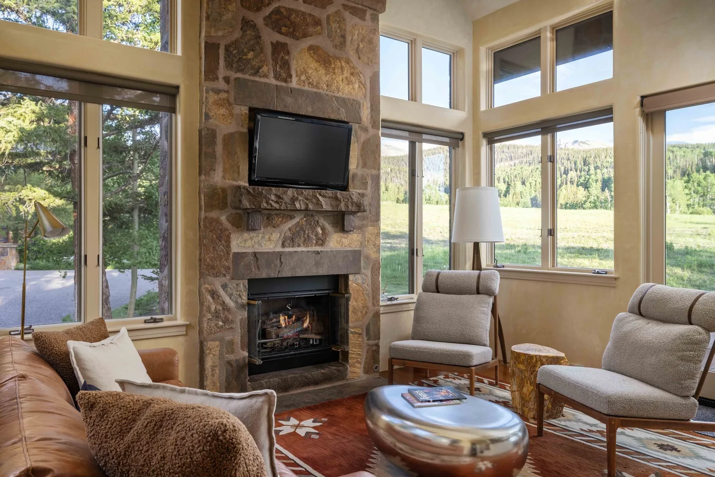 A living room with a stone fireplace, seating, and large windows overlooking greenery.