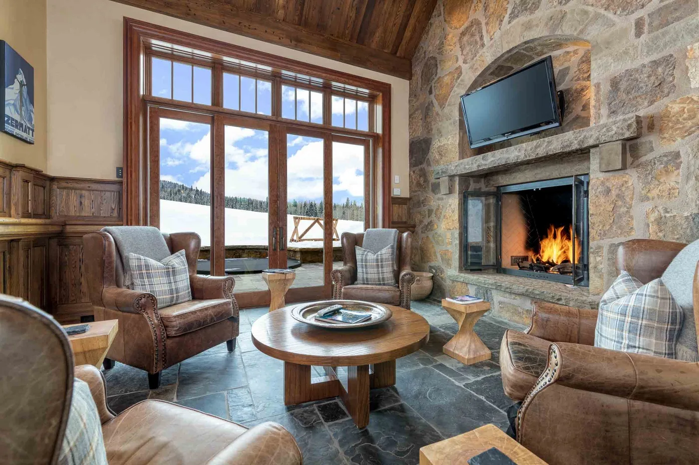 A rustic living room with a stone fireplace, seating, and large windows.