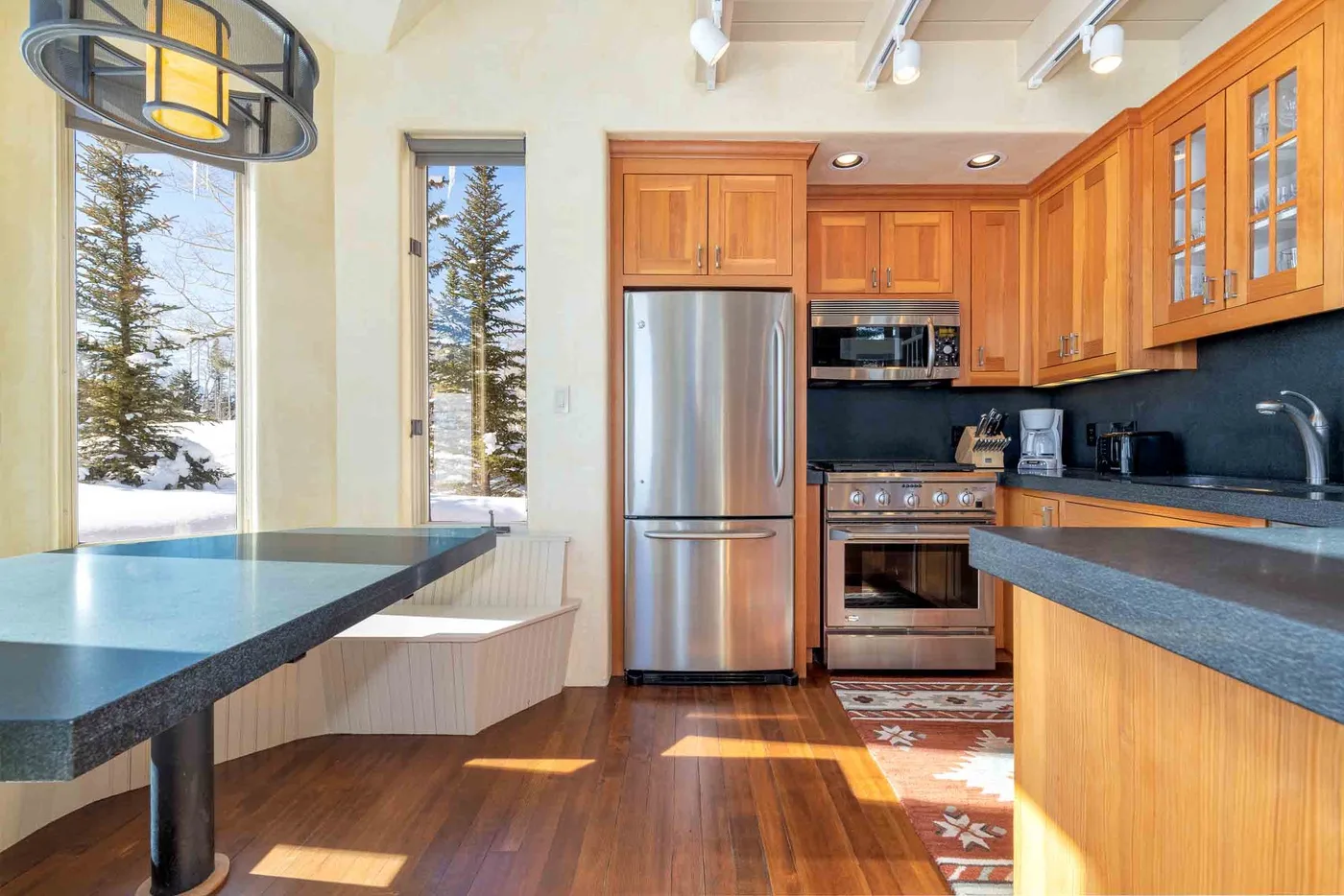 A bright kitchen with wooden cabinets, stainless steel appliances, and a counter beside tall windows.