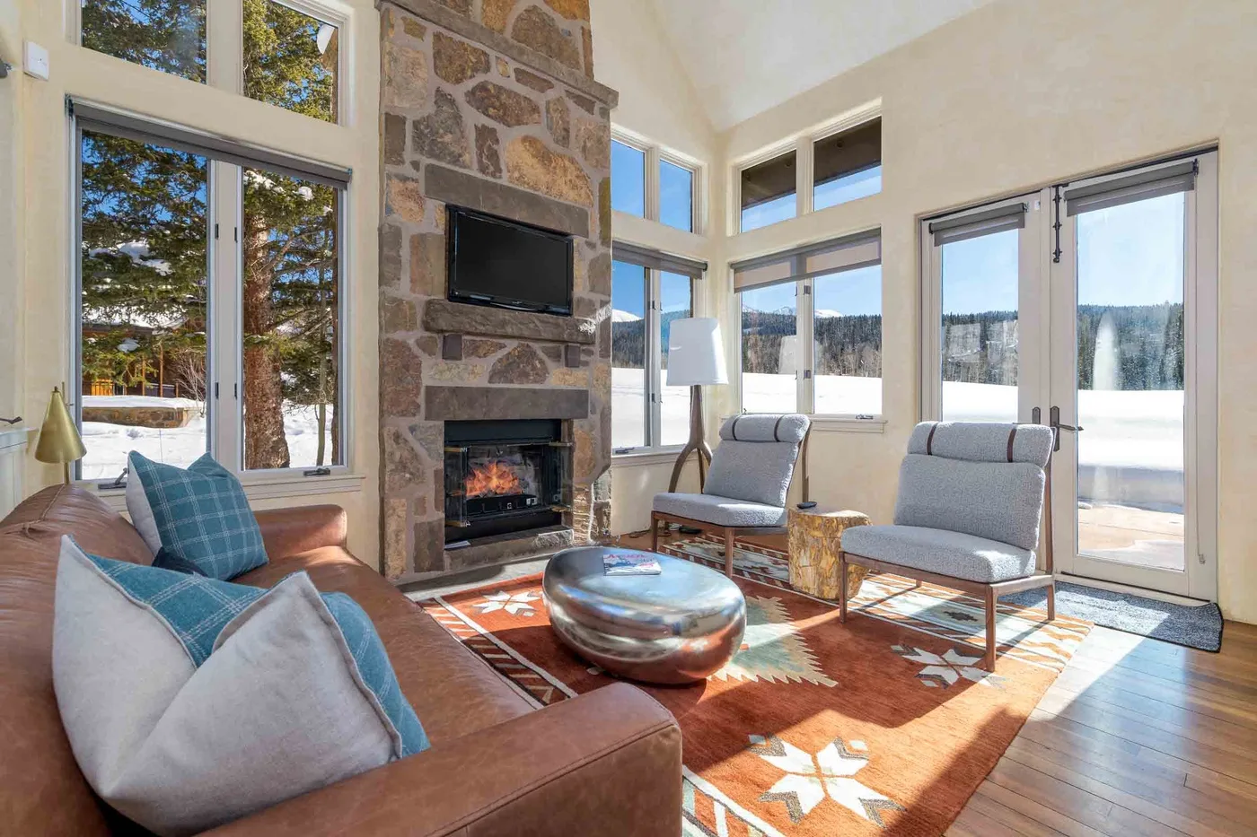 A sunlit living room with a stone fireplace, large windows, and seating.