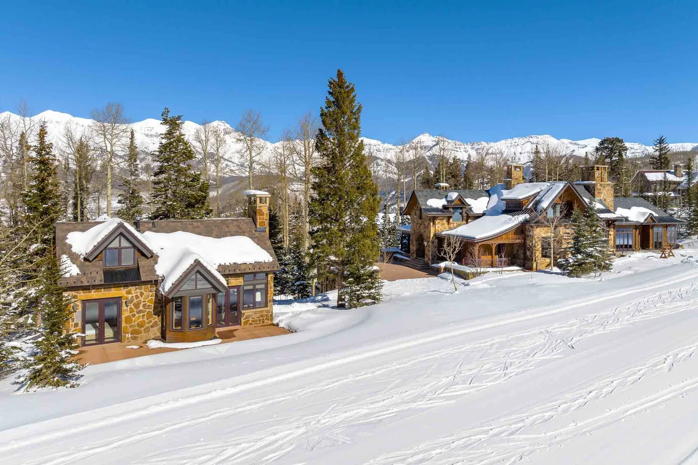 Snow-covered mountain homes sit beside a snowy road beneath a clear blue sky.