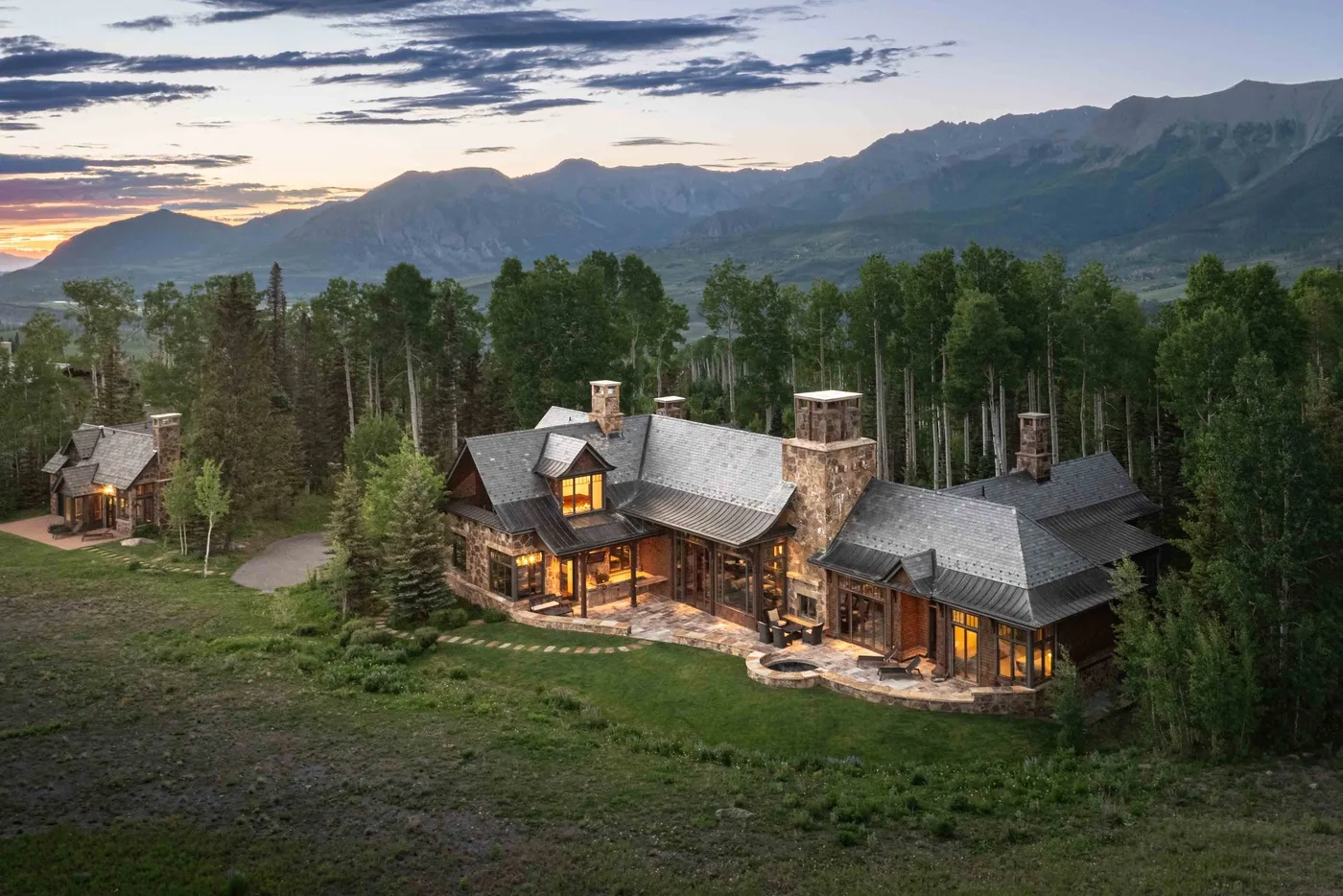 A large lodge sits among trees, with mountains visible beneath a cloudy sunset sky.