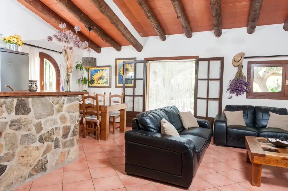 A living and dining area with dark seating, a wooden table, and exposed ceiling beams.