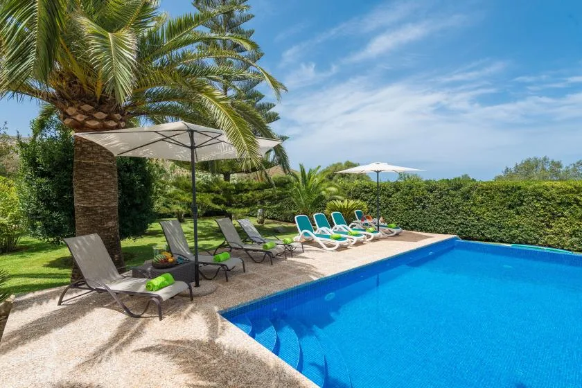 An outdoor swimming pool is bordered by lounge seating, umbrellas, and palm trees.