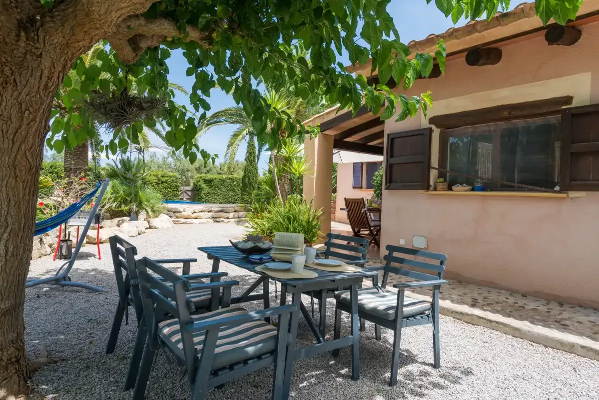 An outdoor dining area with seating sits beside a house under a leafy tree.