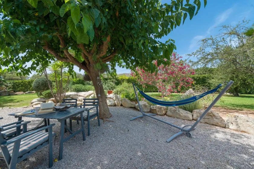 A shaded outdoor seating area with a hammock beneath a leafy tree.