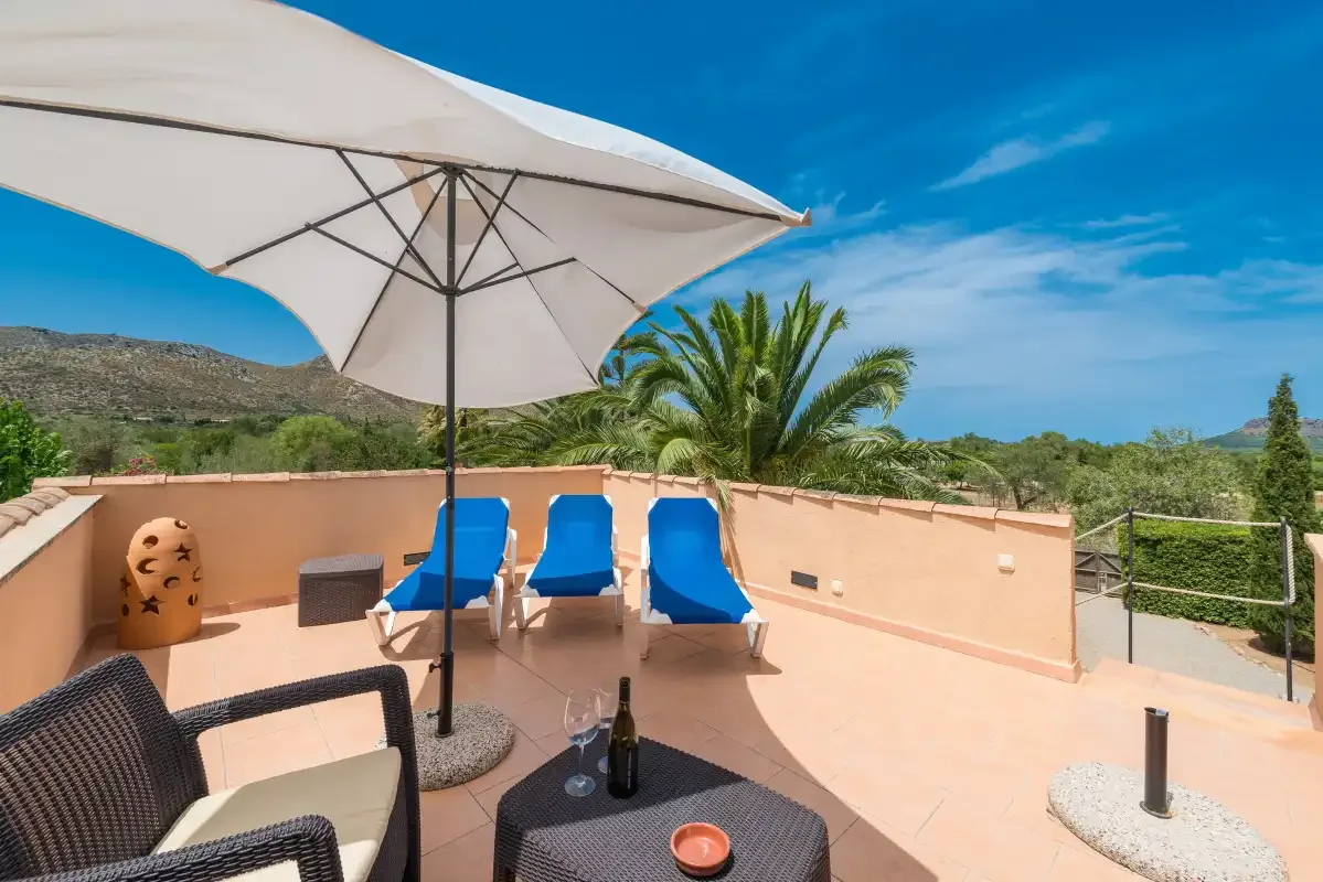 A sunny outdoor terrace has lounge seating, a large umbrella, and views of palm trees.