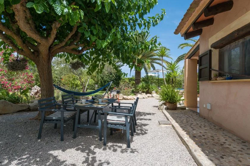 A shaded outdoor patio with seating beneath a leafy tree beside a house.