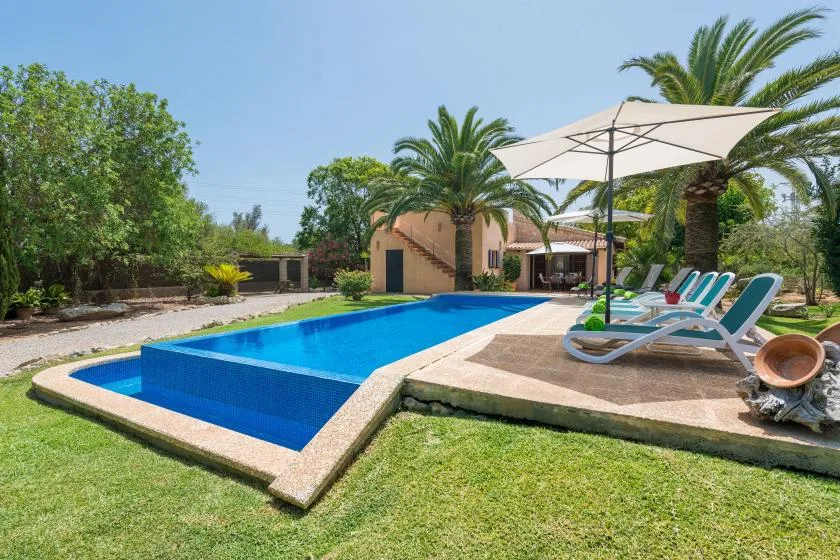 An outdoor swimming pool beside a sunlit patio with loungers and an umbrella.