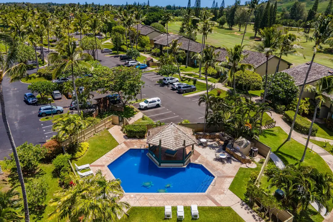 Aerial view of a swimming pool beside a parking area amid tropical greenery.