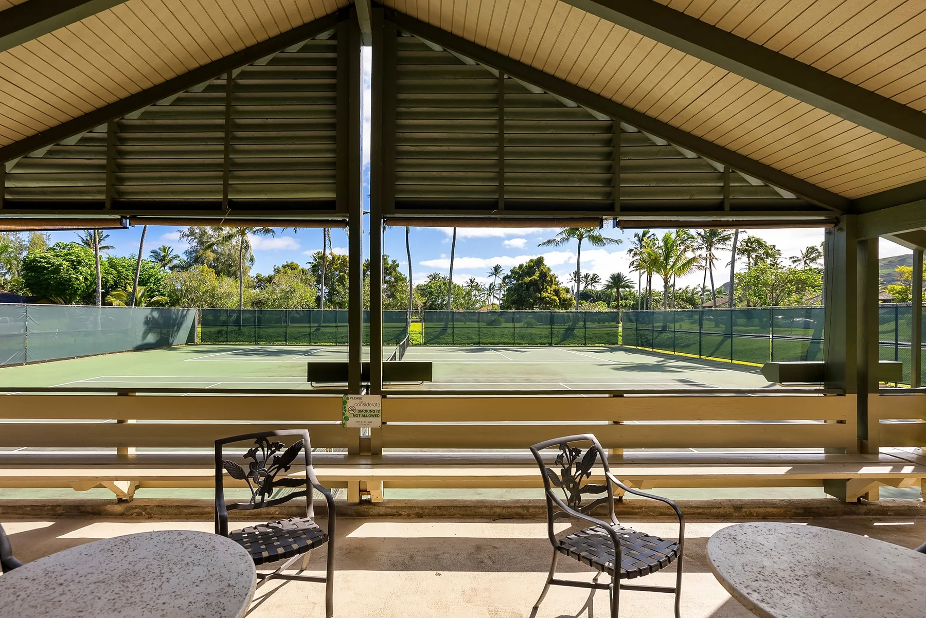 A covered seating area overlooks a fenced tennis court and surrounding trees.