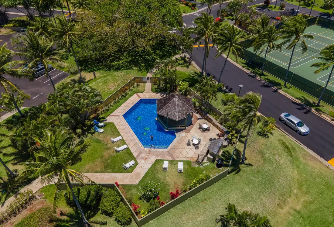 An outdoor swimming pool sits beside a shaded pavilion amid palm trees.