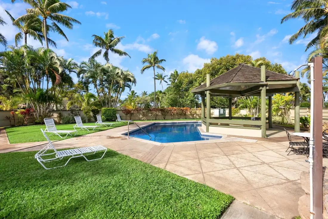 An outdoor swimming pool beside a covered seating area, with lawn and palm trees.