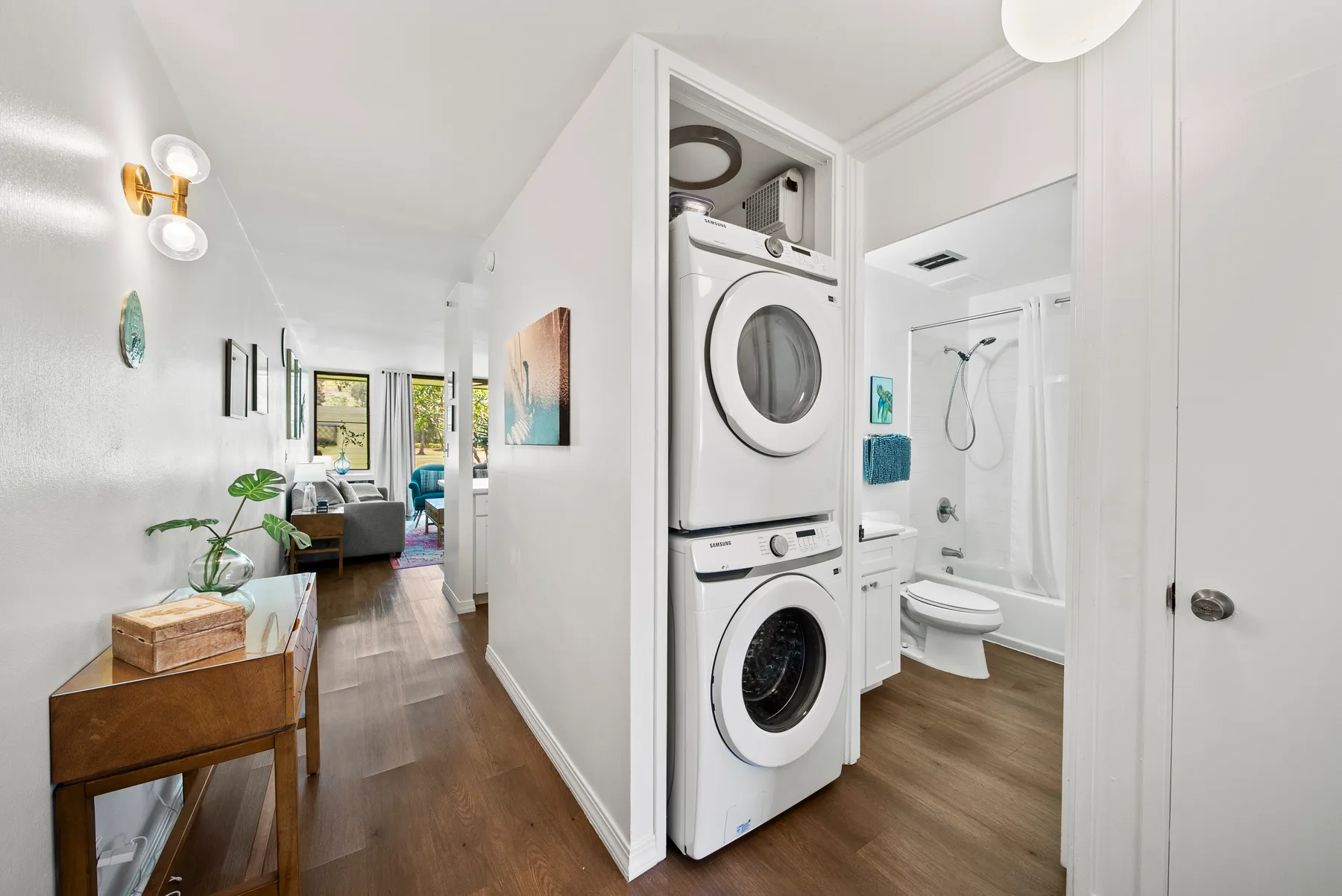 A bright hallway opens onto stacked laundry appliances, a bathroom, and a seating area.