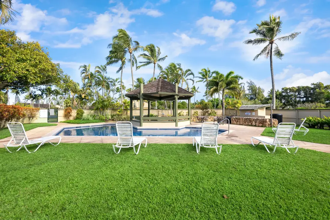 An outdoor swimming pool and gazebo sit amid palm trees and green lawn.