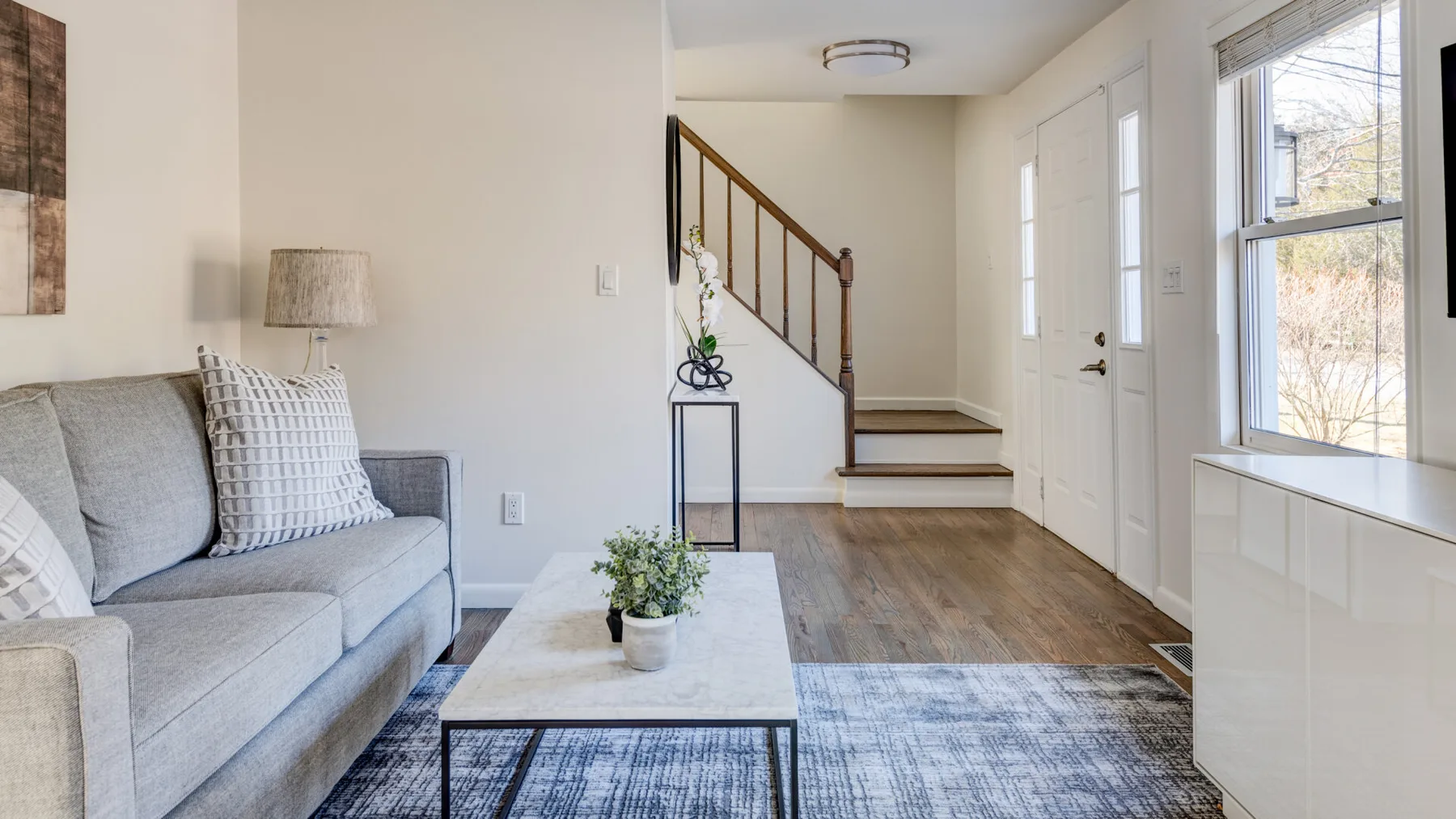 Una sala luminosa con sofá gris, mesa de centro y escalera.