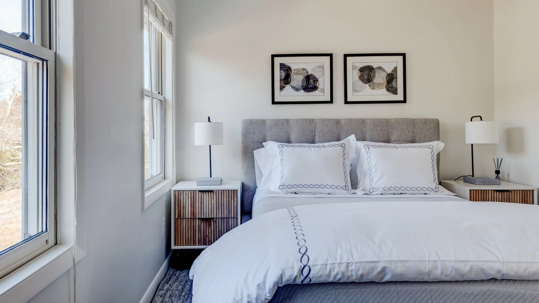 Un dormitorio luminoso con una cama tendida y cuadros enmarcados encima.
