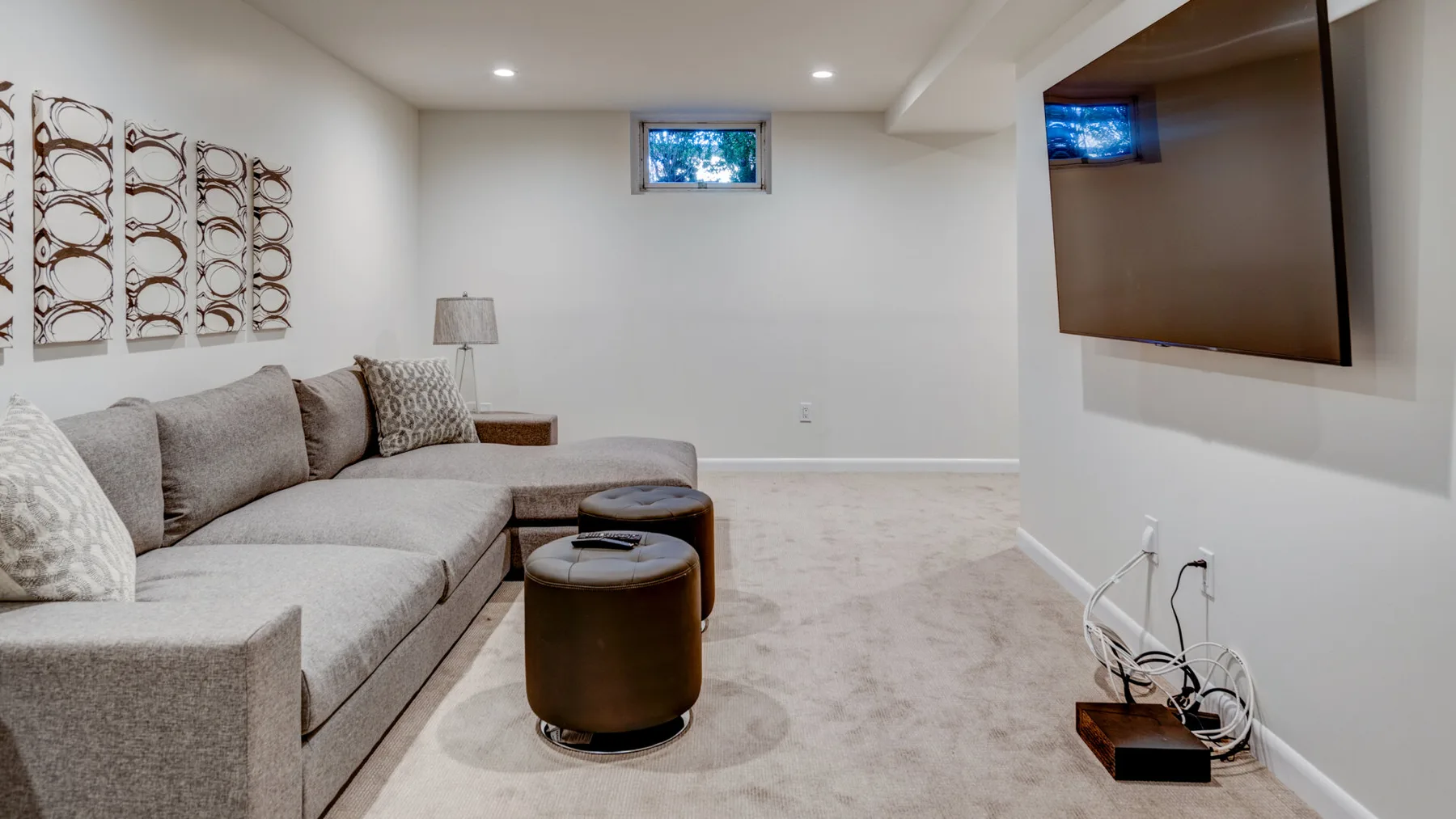Una sala de estar en un sótano con sofá seccional, otomanas y un televisor montado en la pared.