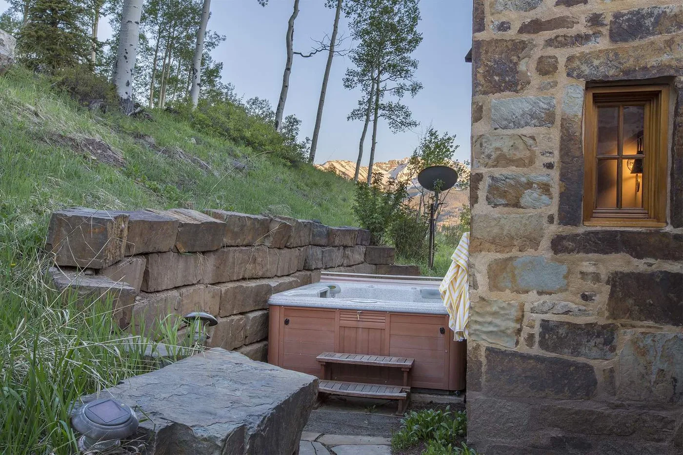 Una bañera de hidromasaje está junto a un muro de piedra en un patio ajardinado.