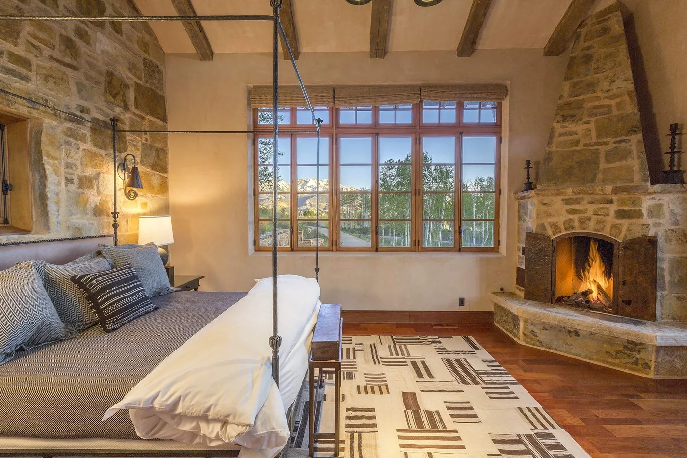 Un dormitorio con cama, alfombra estampada, ventanas grandes y una chimenea de piedra.