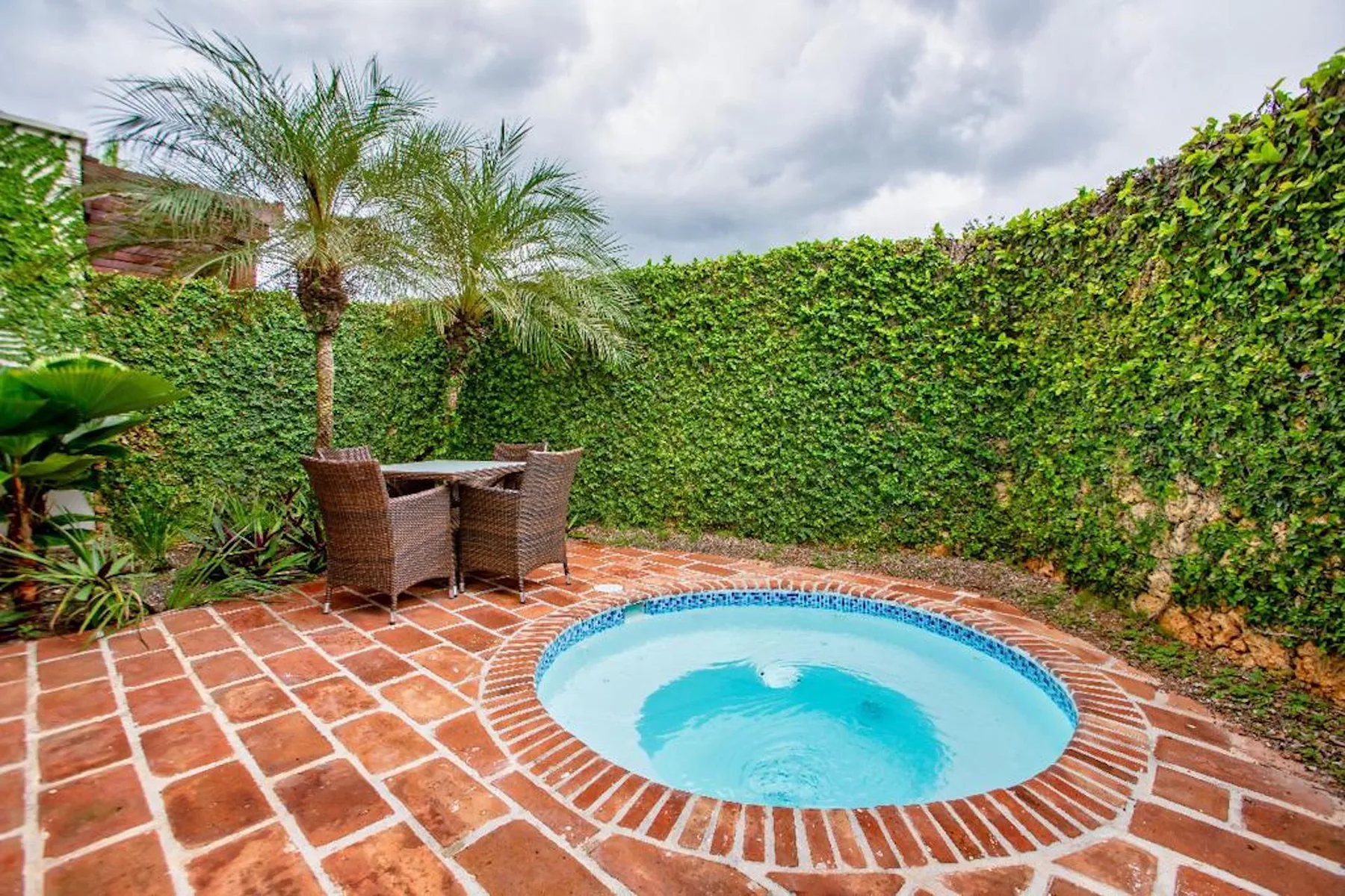 Una pequeña piscina al aire libre está en un patio de ladrillo rodeado de vegetación.