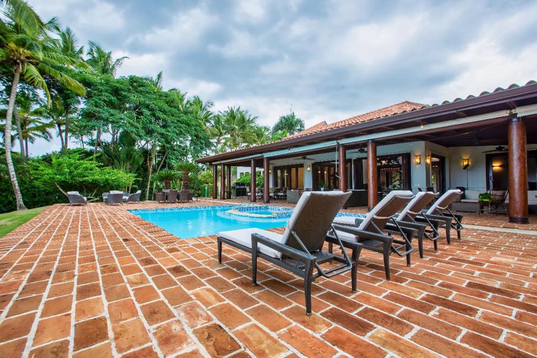 Una piscina y asientos en un patio están junto a un edificio cubierto, rodeados de vegetación tropical.