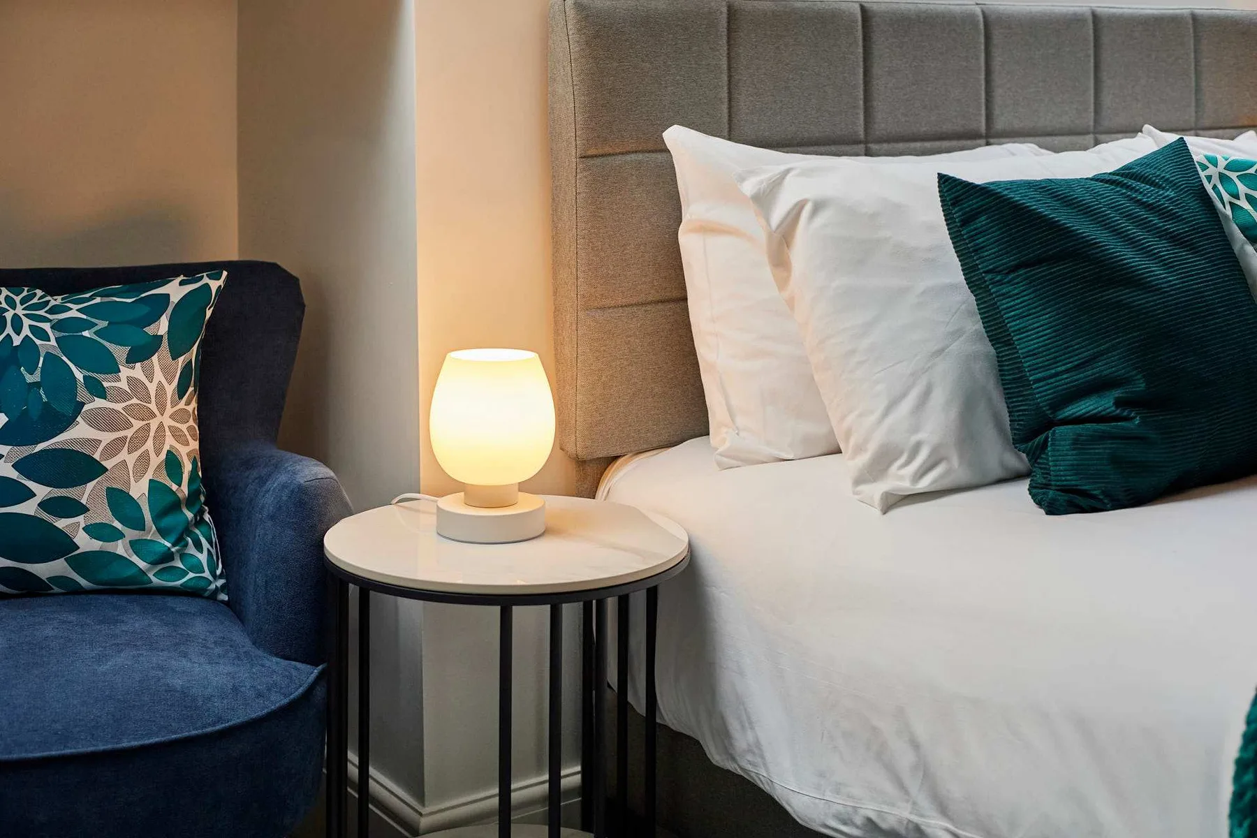 Un dormitorio con una cama tendida, una mesita de noche y una lámpara encendida.
