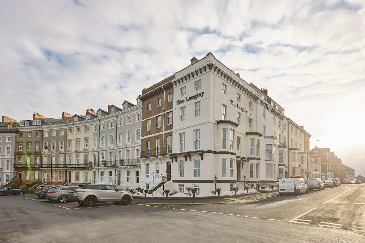 A white corner hotel building stands beside a street lined with parked cars.