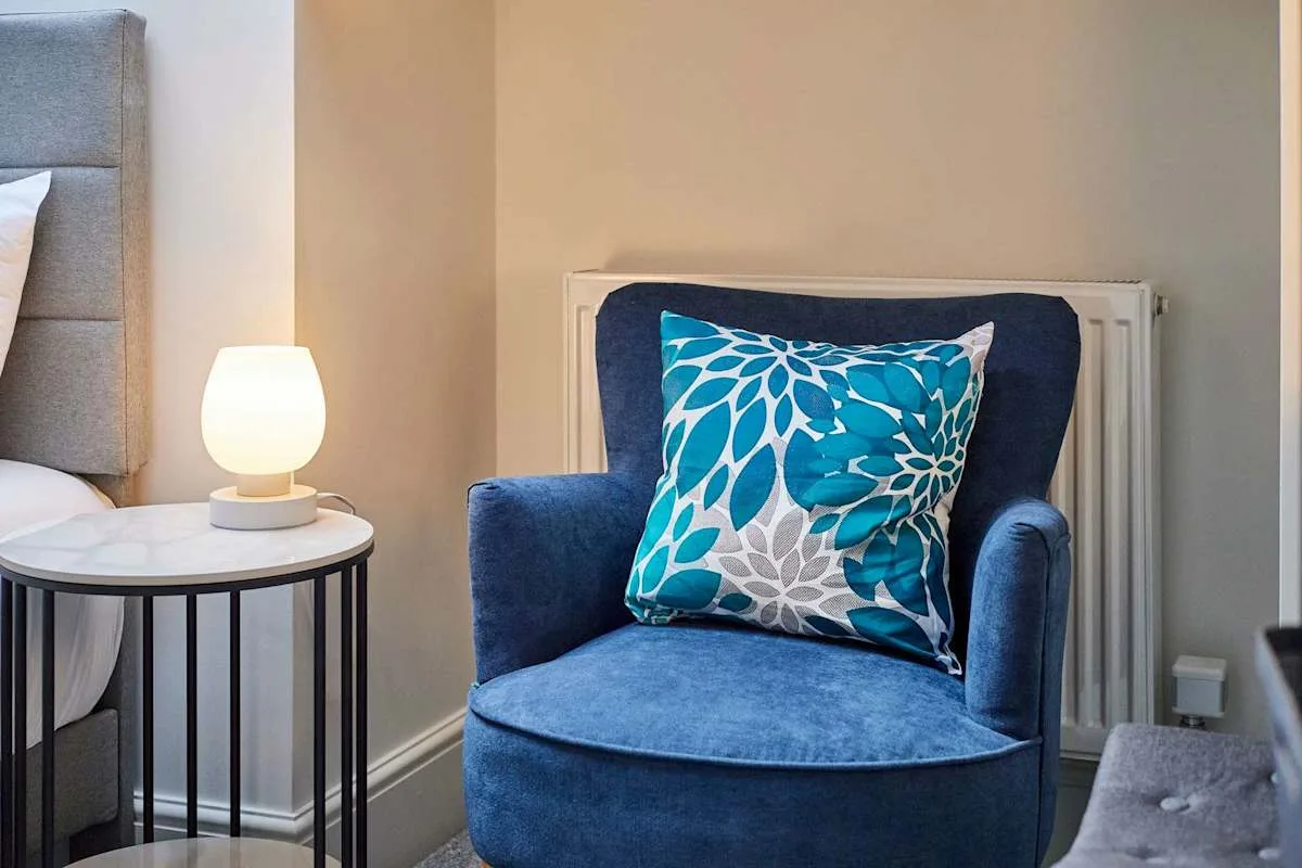 A blue armchair with a patterned cushion sits beside a bed and a lit table lamp.