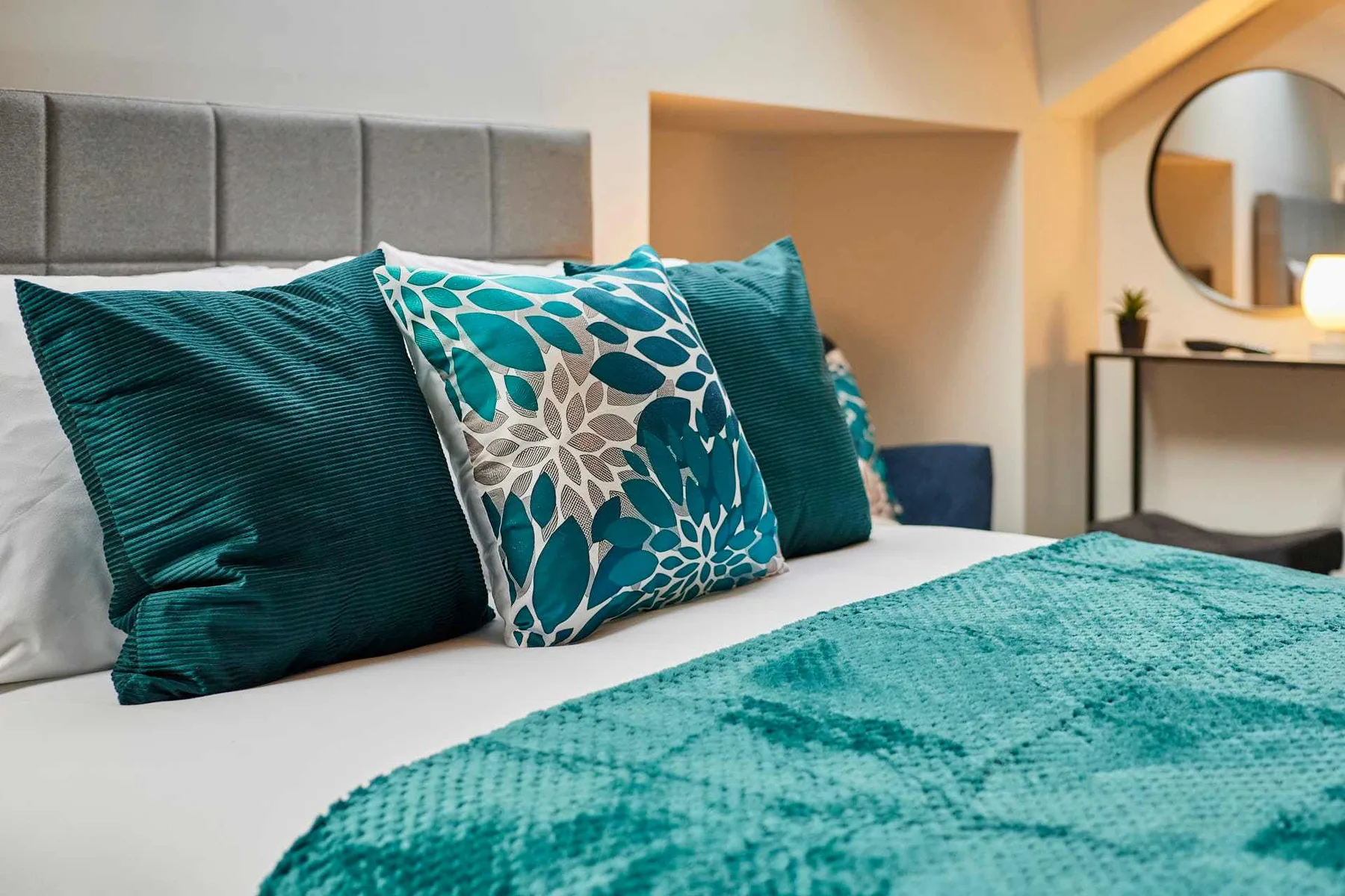A bedroom with a bed dressed in teal bedding and patterned cushions.