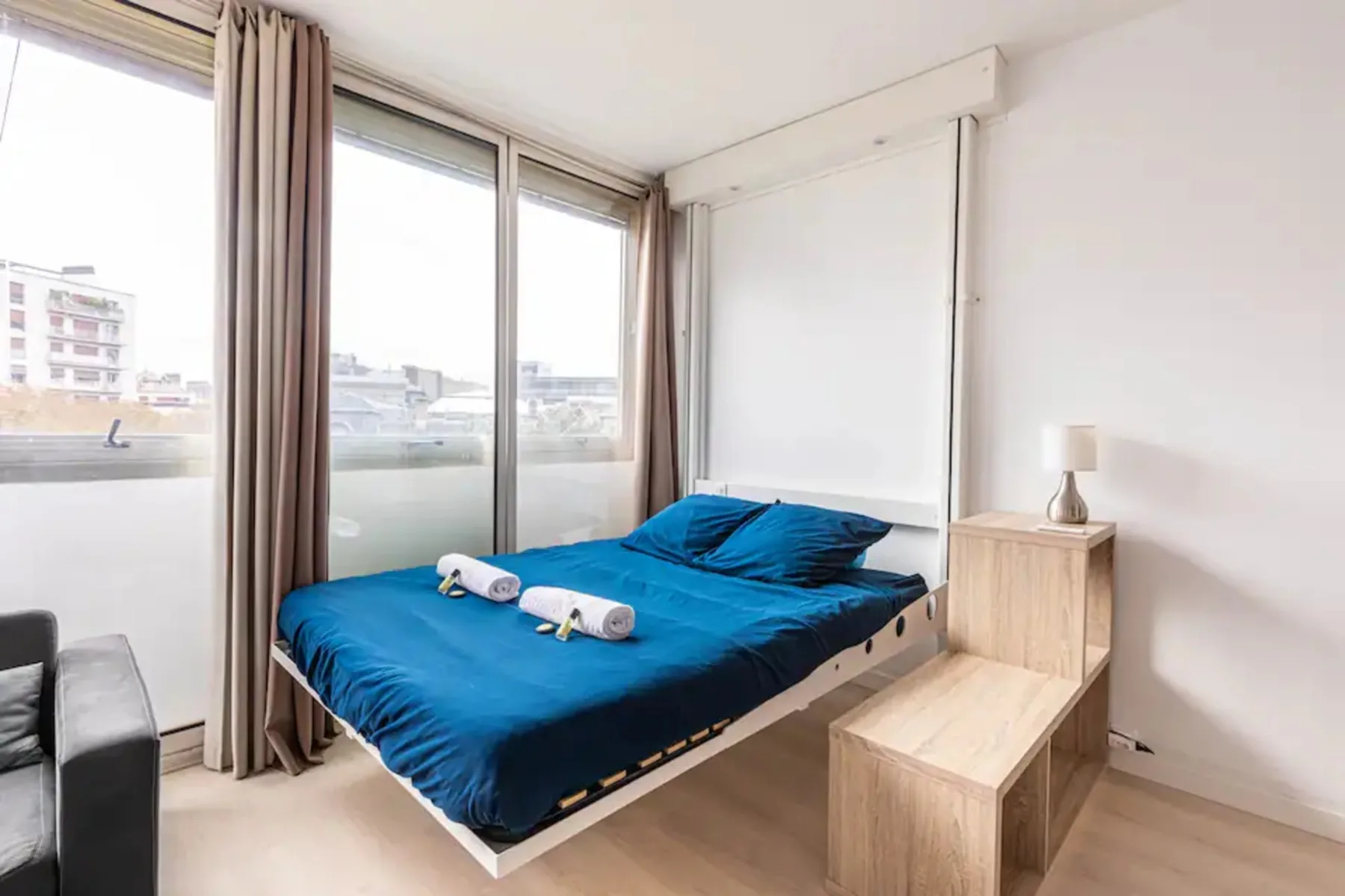 Un dormitorio con una cama azul junto a grandes ventanas y una pequeña mesa de madera.